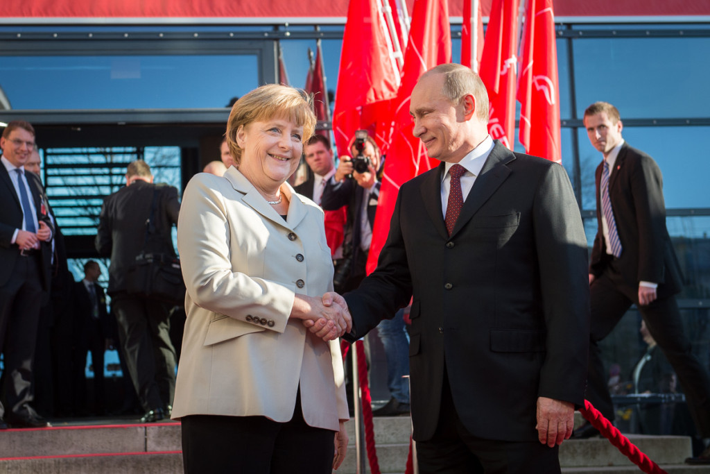 Angela Merkel welcomes Vladimir Putin in front of flagpoles.