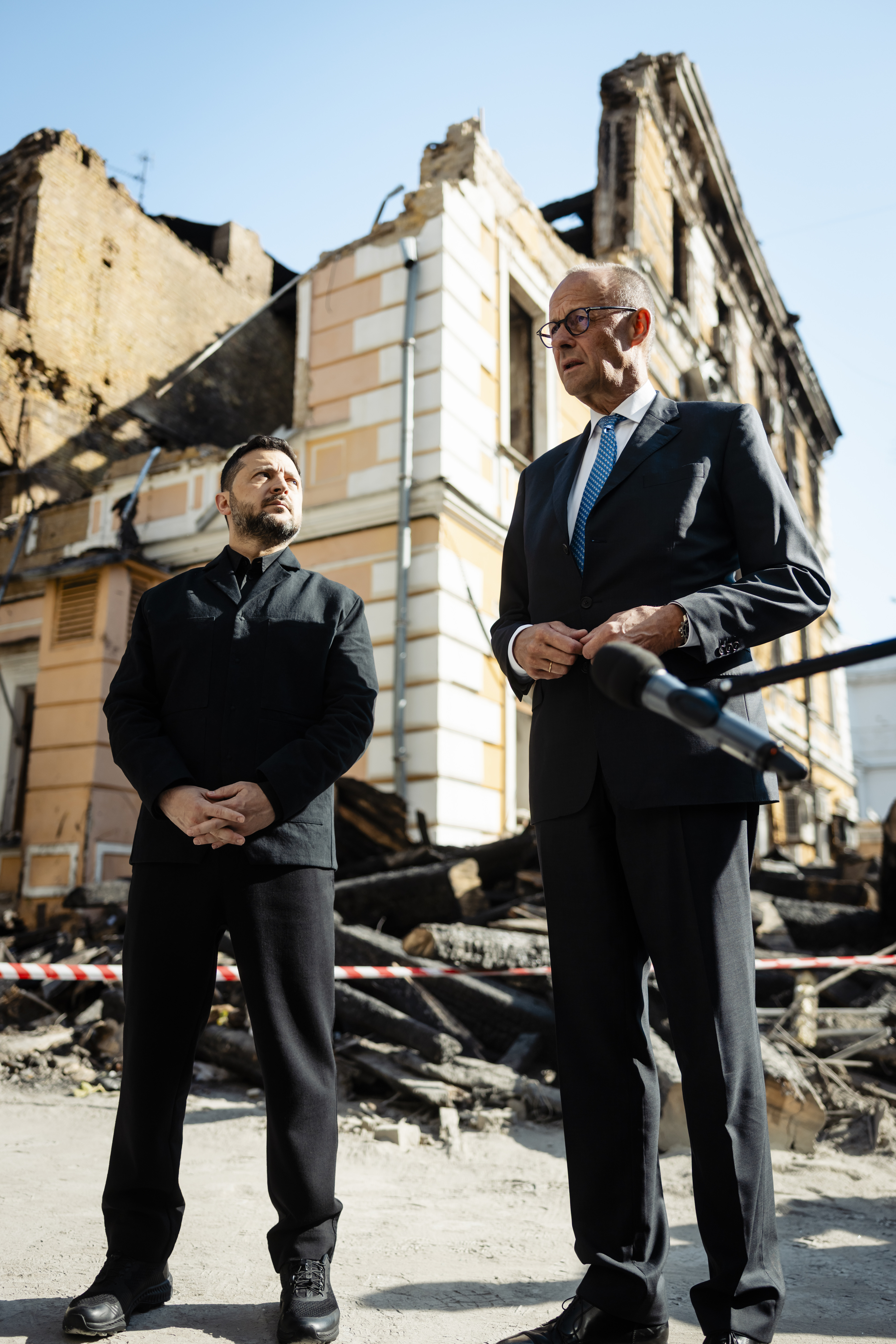 Friedrich Merz und Wolodymyr Selenskyj vor einem zerstörten Haus.