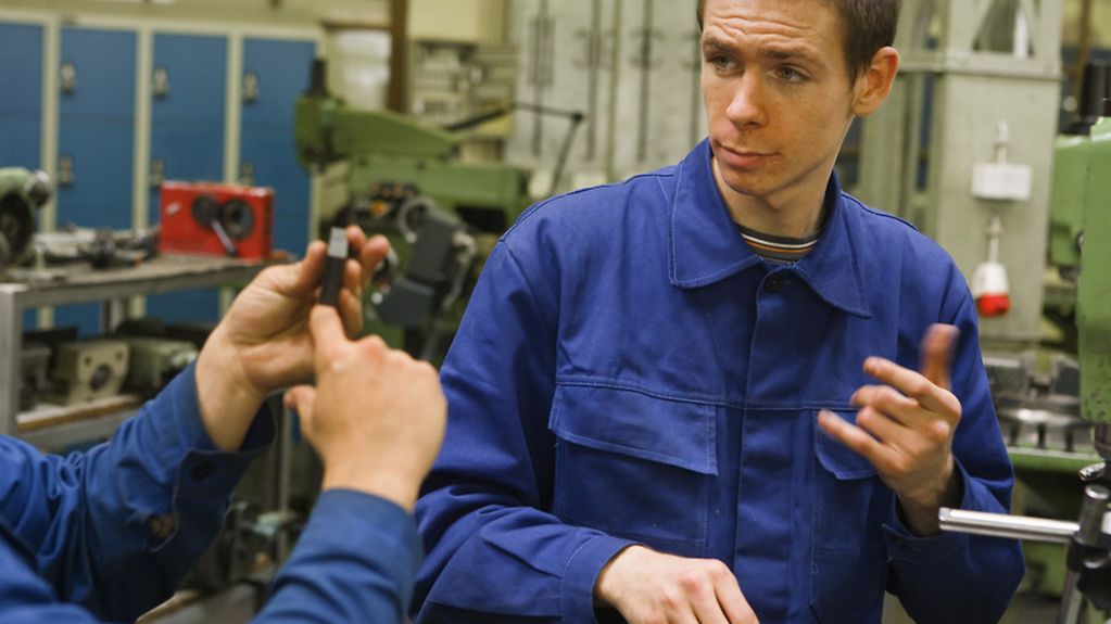 Ein Auszubildender in Arbeitskleidung in einer Metallwerkstatt verständigt sich in Gebärdensprache. 