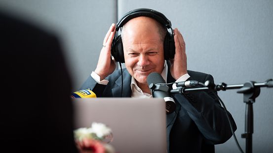 Bundeskanzler Olaf Scholz sitzend mit Kopfhörern.