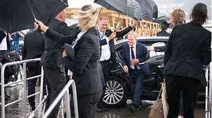 Bundeskanzler Olaf Scholz wird beim Aussteigen aus einem Fahrzeug mit Schirmen vorm Regen geschützt.
