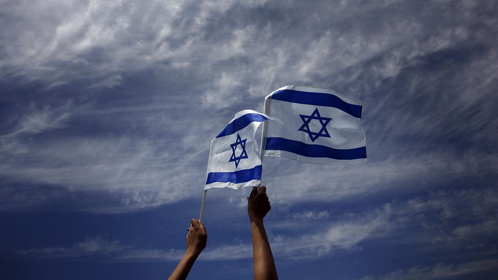 Israelische Flaggen flattern im Wind. Sie werden von zwei Händen in den Himmel gehalten.