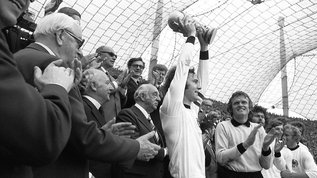 Franz Beckenbauer (mitte) streckt den WM-Pokal in die Luft. Er ist umgeben von Mitspielern und Politikern.