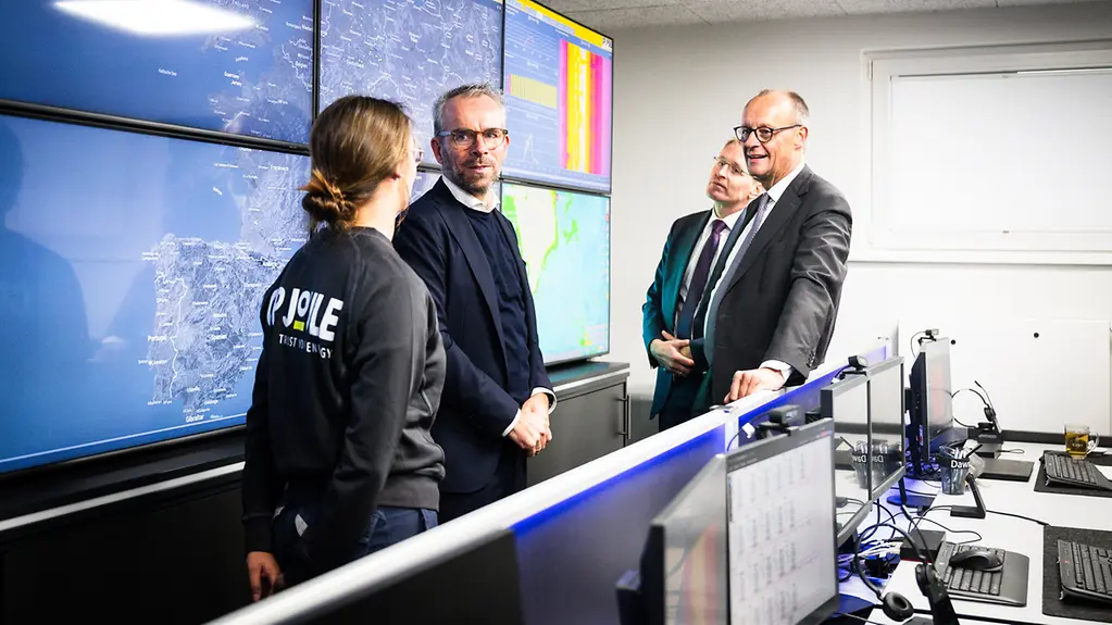Bundeskanzler Merz mit Ministerpräsident Günther und zwei Mitarbeiterinnen von GP Joule vor einer Bildschirmwand der Firma.