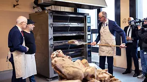 Das Foto zeigt Bundeskanzler Friedrich Merz, der mit Schürze einen Laib Brot in einen Ofen schiebt.
