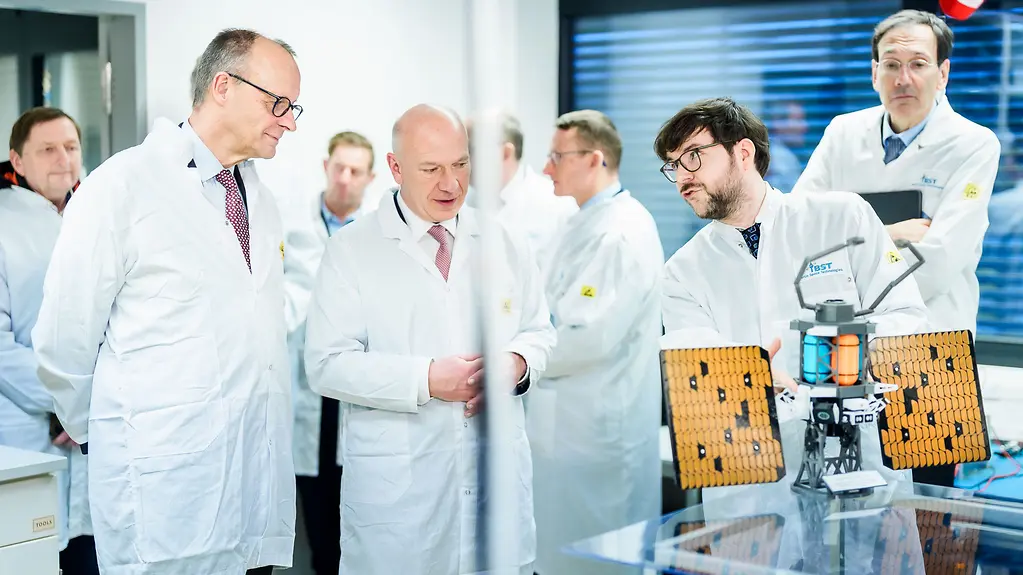 Bundeskanzler Merz, Berlins Regierender Bürgermeister und ein Mitarbeiter von BST vor einem Kleinsatelliten.