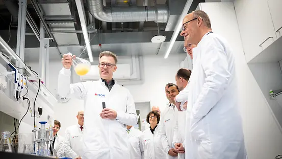 Bundeskanzler Merz im Labor der Firma Tron, gekleidet in einen Laborkittel. Neben ihm Mitarbeiterinnen und Mitarbeiter des Unternehmens.