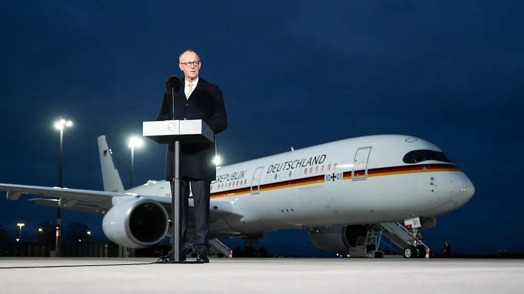 Bundeskanzler Merz auf dem Rollfeld eines Flughafens hinter einem Rednerpult. Im Hintergrund die Regierungsmaschine