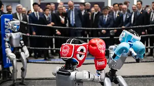 Das Foto zeigt Bundeskanzler Friedrich Merz, der einem Boxkampf von Robotern zuschaut.