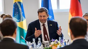 Lars Klingbeil sitzt am Tisch. Im Hintergrund die Flaggen Brasiliens, Deutschlands und der EU.