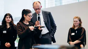 Bundeskanzler Merz im Gespräch mit einer Schülerin. Zwei weitere Schülerinnen schauen zu.