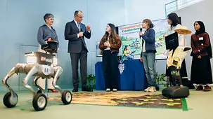 Bundeskanzler Merz im Gespräch mit Schülerinnen. Im Vordergrund stehen zwei kleinere Roboter.