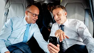 Merz und Macron im Helikopter, beide schauen auf das Smartphone des Kanzlers