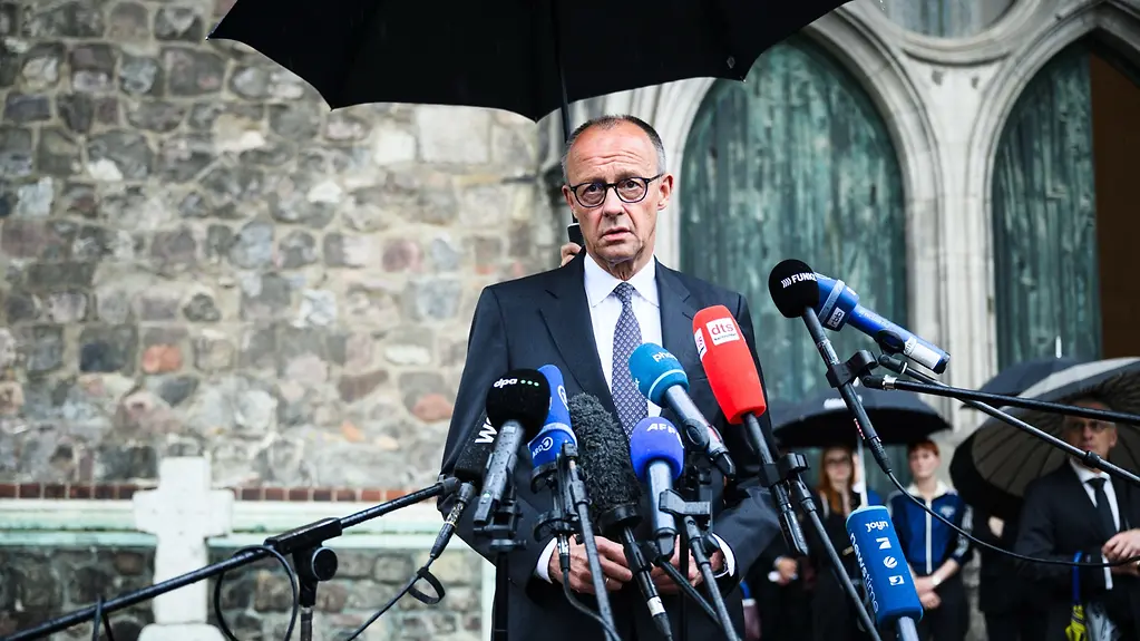 Kanzler Merz beim Statement unterm Regenschirm vor der Kirche.
