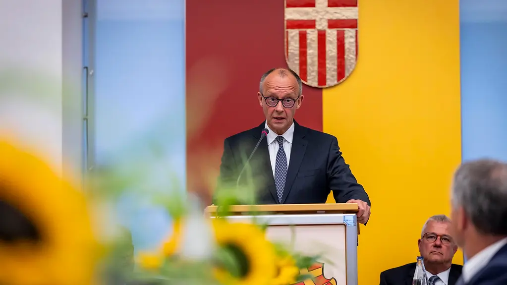 Bundeskanzler Merz vor einem rot-gelben Wappen am Rednerpult. Im Vordergrund unscharfe Sonnenblumen.