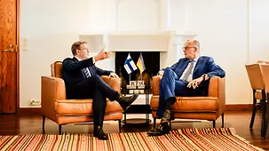 Chancellor Friedrich Merz and Petteri Orpo seated in armchairs in front of a fireplace, deep in conversation.