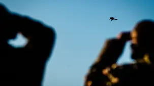 A fighter jet flies in the sky. In the foreground, two figures can be seen in silhouette, watching the aeroplane through binoculars.