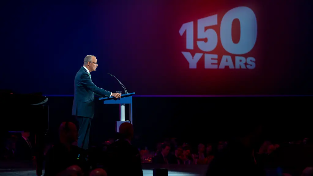 Bundeskanzler Merz steht auf einer Bühne, im Hintergrund der Schriftzug "150 Years".