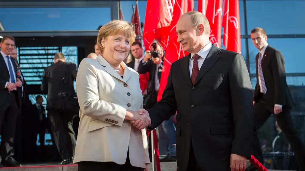Angela Merkel welcomes Vladimir Putin in front of flagpoles.
