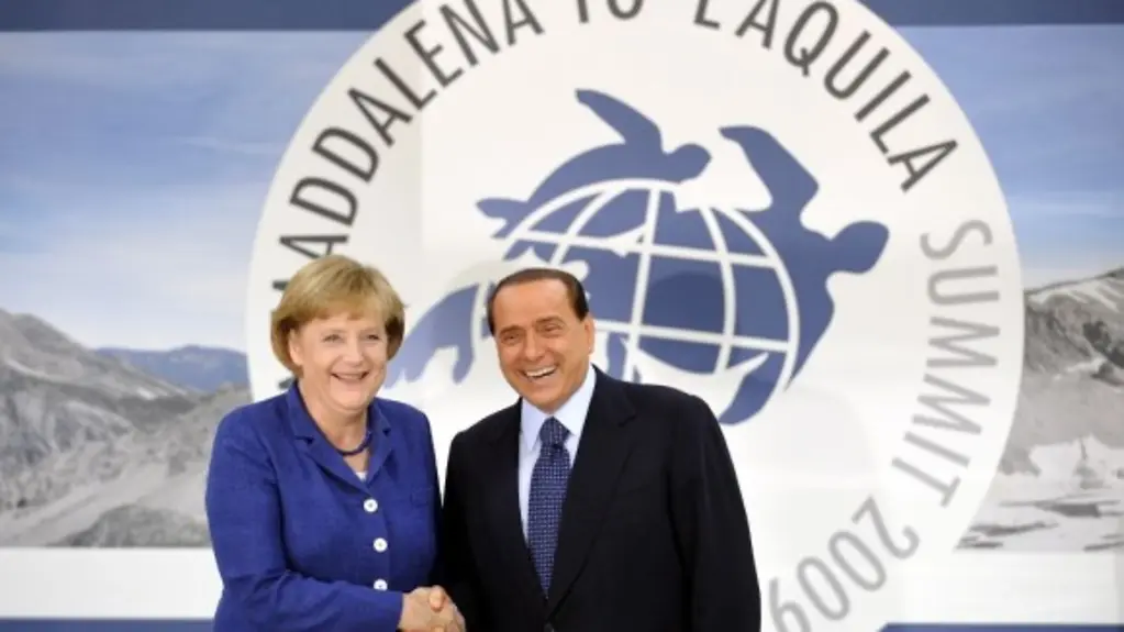 Silvio Berlusconi and Angela Merkel shake hands against a backdrop of the G8 flags